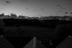 Monochrome, early morning sunrise, trees in the background, meadow and a roof in the foreground.