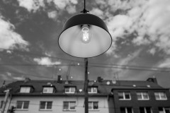 A lamp, with bright sky with clouds and some houses in the background