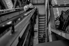 Two people are seen from above, going down a narrow staircase in a massive industrial structure