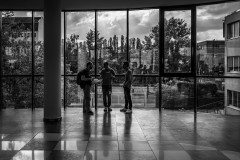 Silhouetted figures, engaged in conversation, stand in front of a glass wall, a cloudy sky and a line of trees behind them outside