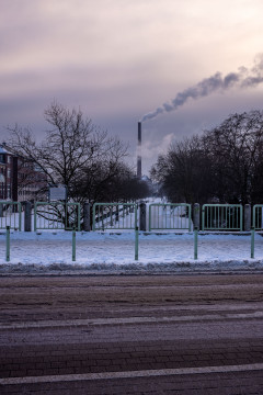 Street and snow covered sidewalk in the foreground, trees and a large chimney with smoke plume in the background