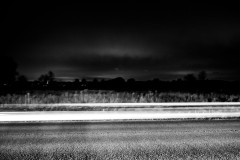 Dark street, with the lights of a car streaking by, dark field and sky in the background