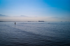 Calm, blue ocean in the foreground, people bathing in the middle ground and two large ships docked and misty mountains far in the background