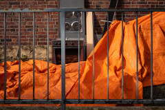 A bright orange tarp, half covering bulky waste, eg. an oven, a handrail in the foreground, brick wall in the back