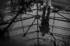 Silhouetted reflection in a puddle of water of someone taking a picture, the persons shoes are also seen, standing in the puddle