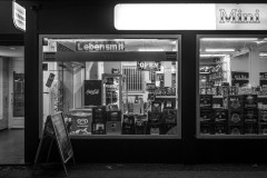 Monochrome image of the front of a convenience store (mini market) after dark being illuminated from inside, with beer crates in the window