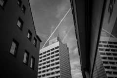 Monochrome image: Two buildings frame a highrise building, which is also reflected in the window of the building to the right; two condensation trails are seen in the sky above the highrise