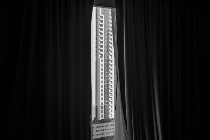 Monochrome image of a view through a window, with curtains half drawn, looking at a highrise building