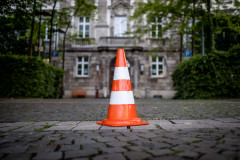 A orange traffic cone in the center of the frame, with a building and some trees in the background