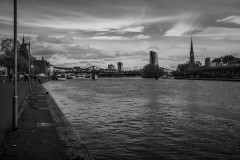 A black and white urban landscape photograph taken along a river. A walkway or promenade runs along the riverbank in the foreground. The city skyline features a mix of modern office towers and historic church spires with pointed steeples. A pedestrian bridge with metal framework spans across the river. Dramatic clouds fill the sky above the cityscape, and the river's surface shows rippling patterns. A few people can be seen walking along the riverside path.