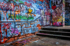 The image shows a heavily graffitied wall, with various colors, shapes, and text scrawled across the surface. There are stairs leading up to the wall, and the overall scene looks like it is in an urban, outdoor setting.