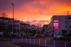 This image depicts an urban sunrise scene with a vibrant sky. The sky is ablaze with warm hues of orange, red, and pink, creating a dramatic backdrop. There are buildings, streetlights, and a pedestrian crossing the street.