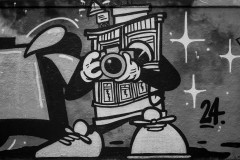 This monochrome image shows a graffiti-style artwork on a wall; it features abstract shapes, geometric patterns, and a stylized cartoon-like character which is looking through a camera