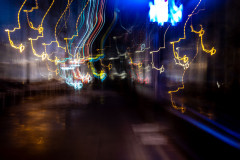 Abstract, long-exposure photograph taken early in the morning, capturing streaks of light in various colors. The predominant colors are yellow, blue, and white light trails that create curved and zigzagging patterns against a dark background. The effect creates a dynamic, almost chaotic composition that suggests movement through an urban environment.
