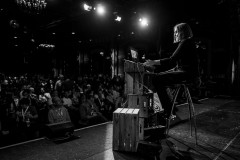 Léoni Watson on stage at beyond tellerrand 2025 in Düsseldorf, seated on a high stool, while working her laptop; the black and white image shows her in profile, illuminated by stage lights, with the audience visible in the darkened background
